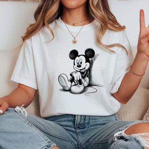 K&ouml;nnte beinhalten: Wei&szlig;es T-Shirt mit einer Schwarz-Wei&szlig;-Illustration von Mickey Mouse, der an einer Wand sitzt und sich anlehnt. Die Zeichentrickfigur tr&auml;gt wei&szlig;e Schuhe und Handschuhe. Das Shirt besteht aus einem weichen Material.