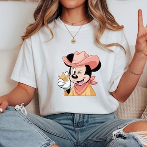 May include: White t-shirt featuring a cartoon of a winking mouse wearing a pink cowboy hat, a pink bandana, and a sheriff's badge. The mouse is holding the badge in its gloved hand. The shirt is paired with blue jeans.