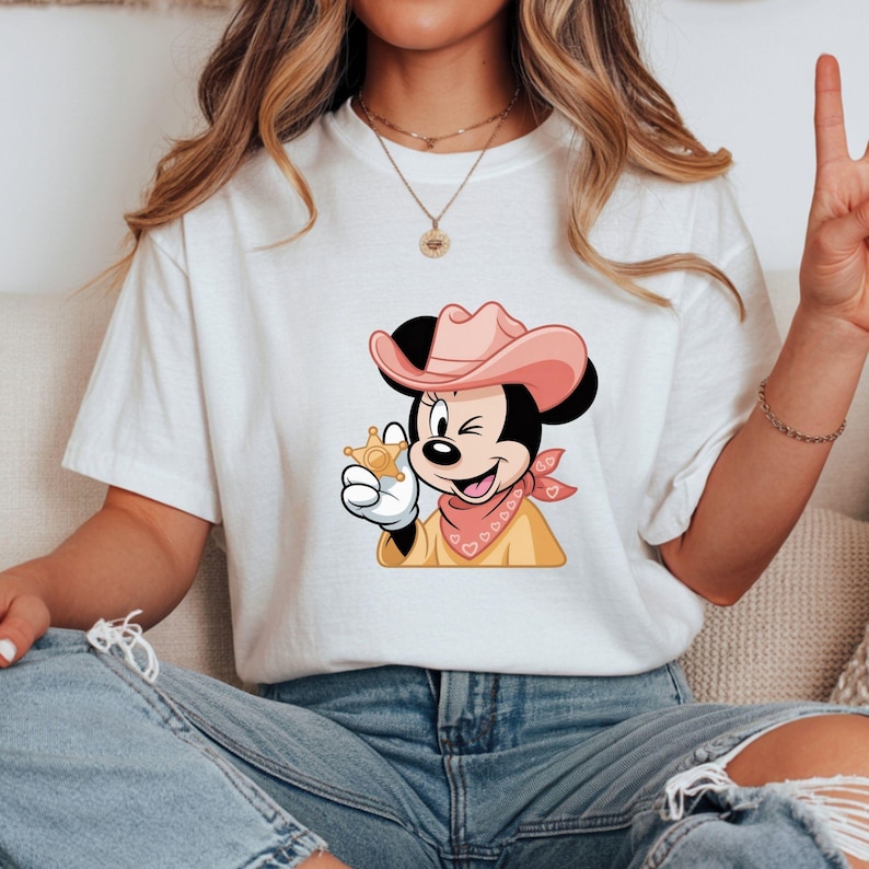 May include: White t-shirt featuring a cartoon of a winking mouse wearing a pink cowboy hat, a pink bandana, and a sheriff's badge. The mouse is holding the badge in its gloved hand. The shirt is paired with blue jeans.