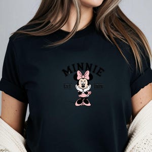 Może przedstawiać: Czarny t-shirt z grafiką Myszki Minnie. Projekt zawiera słowo "MINNIE" w łukowatych literach nad kresk&oacute;wkowym wizerunkiem Myszki Minnie w r&oacute;żowej kokardce i sukience. Tekst "EST 1928" znajduje się poniżej.