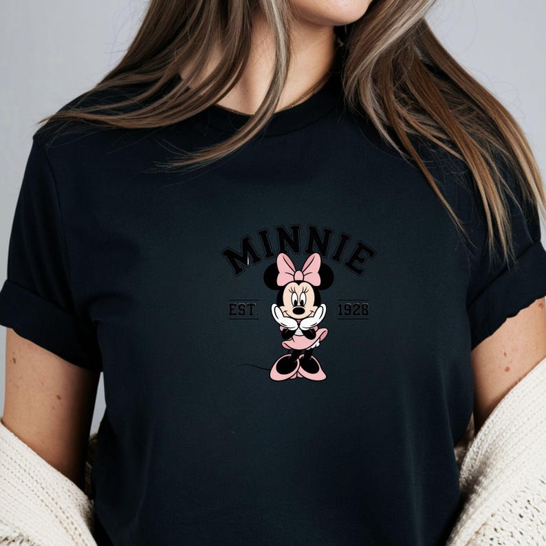 Może przedstawiać: Czarny t-shirt z grafiką Myszki Minnie. Projekt zawiera słowo "MINNIE" w łukowatych literach nad kresk&oacute;wkowym wizerunkiem Myszki Minnie w r&oacute;żowej kokardce i sukience. Tekst "EST 1928" znajduje się poniżej.