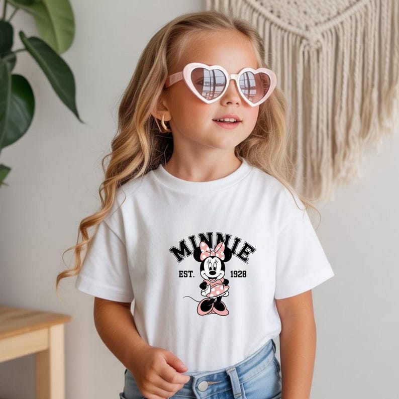 Może przedstawiać: Biała koszulka z grafiką Myszki Minnie. Grafika zawiera słowo "MINNIE" w czarnym, łukowatym kroju pisma, obraz Myszki Minnie oraz tekst "EST. 1928". Dziewczynka ma na sobie r&oacute;żowe okulary przeciwsłoneczne w kształcie serca.
