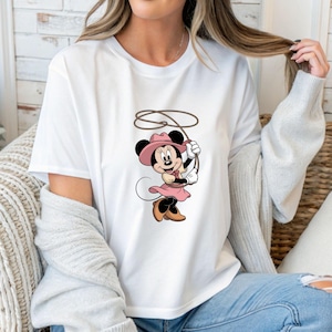 May include: White t-shirt featuring a cartoon Minnie Mouse dressed as a cowgirl, wearing a pink hat and dress, holding a rope. The graphic is centered on the shirt.