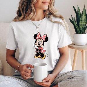 May include: White t-shirt featuring a Minnie Mouse graphic. The cartoon character is depicted in a classic pose, wearing a pink polka-dot bow, dress, and shoes. The shirt is a casual style, suitable for everyday wear.