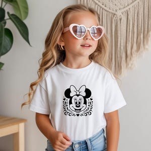 Może przedstawiać: Biała koszulka z czarno-białą grafiką Myszki Minnie. Projekt zawiera twarz Minnie, kokardę i dekoracyjne elementy kwiatowe. Koszulka ma okrągły dekolt.