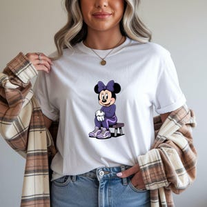 May include: White t-shirt featuring a cartoon Minnie Mouse character in a purple tracksuit and sneakers. Minnie Mouse is seated on a small wooden bench. The shirt is a classic crew neck style.
