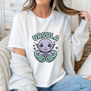 以下が含まれることがあります： Ursulaの漫画イラストが描かれた白いTシャツ。「URSULA」という名前がキャラクターの上に水色で書かれています。キャラクターは紫で、ネックレスとティールアクセントが付いています。4つの小さな星がキャラクターを囲んでいます。