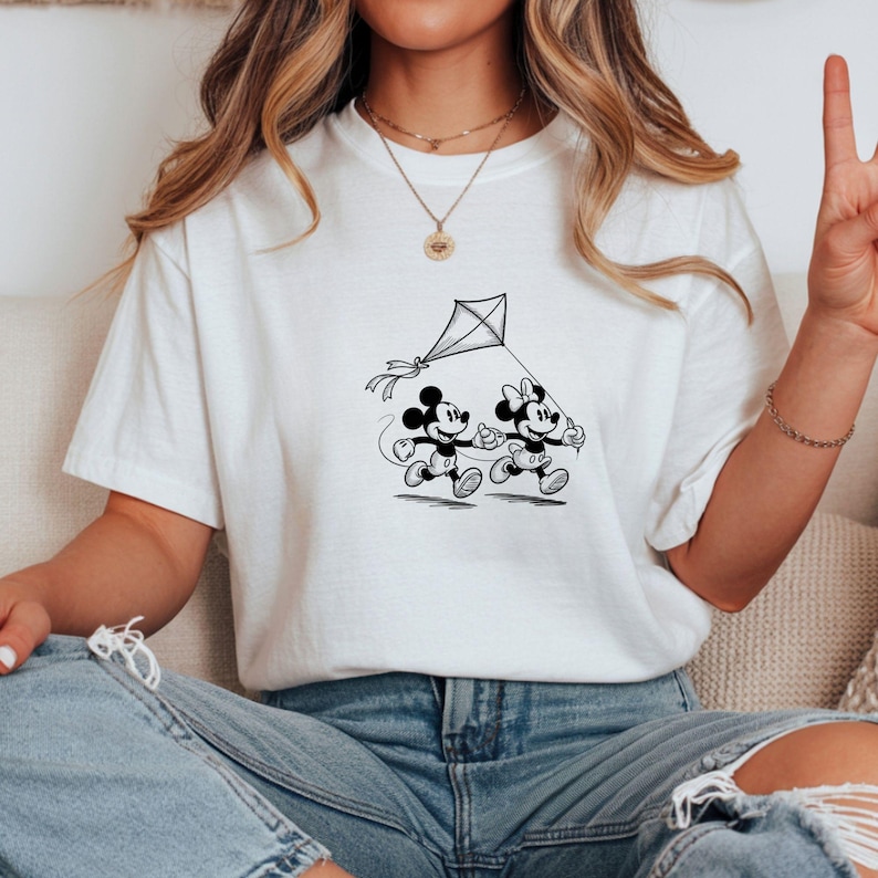 Puede incluir: Camiseta blanca con un gr&aacute;fico en blanco y negro de Mickey y Minnie Mouse corriendo con una cometa. Los personajes de dibujos animados sonr&iacute;en y la cometa est&aacute; en el aire. La camiseta es de material suave.