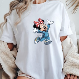 May include: White t-shirt featuring a cartoon illustration of Minnie Mouse in a blue tracksuit and yellow sneakers. Minnie Mouse is wearing a red bow with white polka dots. The graphic is centered on the front of the shirt.