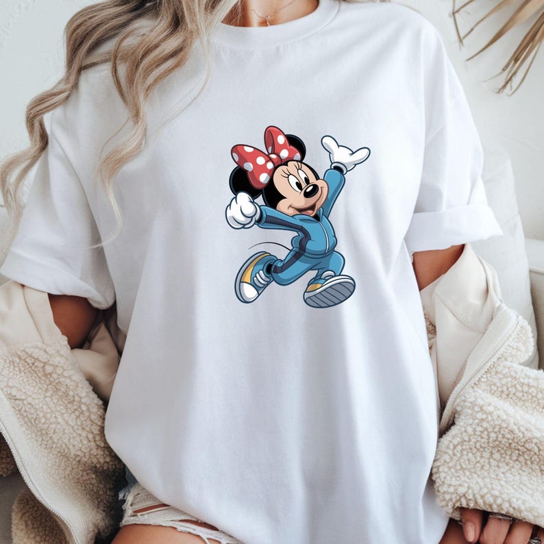 May include: White t-shirt featuring a cartoon illustration of Minnie Mouse in a blue tracksuit and yellow sneakers. Minnie Mouse is wearing a red bow with white polka dots. The graphic is centered on the front of the shirt.