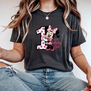 May include: Dark gray t-shirt featuring a graphic of Minnie Mouse with a pink polka-dotted number 1 and the text "of the Birthday Girl". Minnie Mouse is wearing a pink bow and dress. The shirt is a comfortable, casual style.