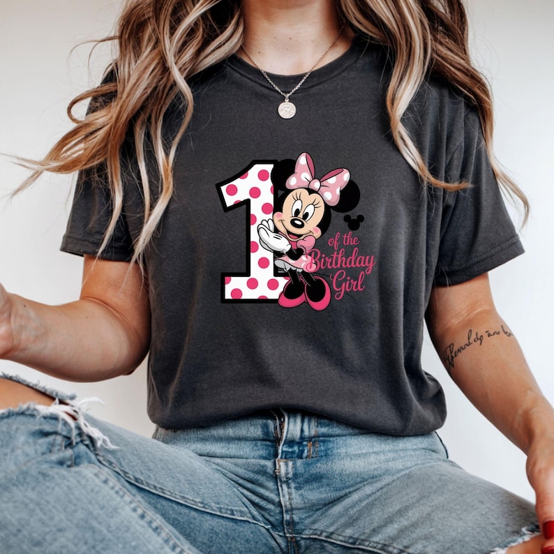 May include: Dark gray t-shirt featuring a graphic of Minnie Mouse with a pink polka-dotted number 1 and the text "of the Birthday Girl". Minnie Mouse is wearing a pink bow and dress. The shirt is a comfortable, casual style.