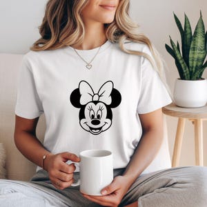May include: White t-shirt featuring a black and white graphic of Minnie Mouse's face. The design includes her signature bow and smiling expression. The shirt is a classic crew neck style.