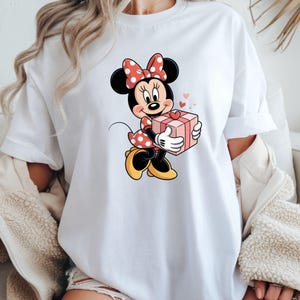 Op de afbeelding: Wit T-shirt met een cartoonontwerp van Minnie Mouse die een roze cadeauverpakking met een rood hart vasthoudt. Minnie draagt een rode polkadot jurk en strik. Kleine roze hartjes zweven rond het personage.