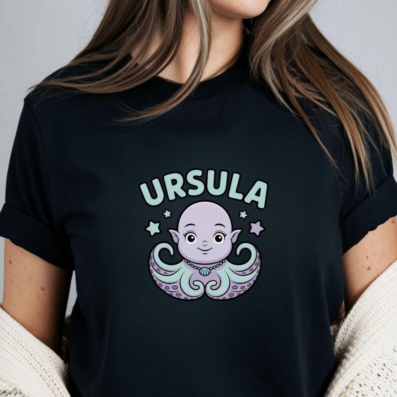 以下が含まれることがあります： 黒いTシャツ。漫画のタコのデザイン。タコは紫とティール色で、真珠のネックレスと星のアクセントが付いています。「URSULA」という名前がタコの上にあります。デザインはTシャツの中央に配置されています。