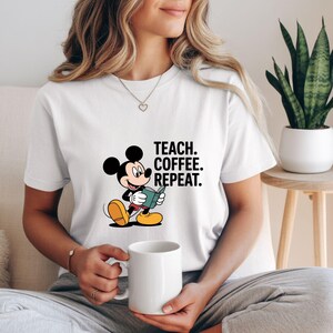 May include: White t-shirt with a Mickey Mouse graphic reading a book, and the text "TEACH. COFFEE. REPEAT." in black. The cartoon character has black ears, yellow shoes, and red shorts. A white mug is held in the person's hands.