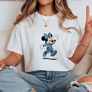 May include: White t-shirt featuring a cartoon Minnie Mouse character in a blue outfit, including a bow, jacket, and shoes. The character is depicted in a running pose with a waving hand. The shirt is worn by a person.