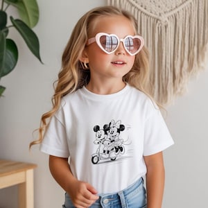 Puede incluir: Una camiseta blanca con una ilustraci&oacute;n en blanco y negro de Mickey y Minnie Mouse en un scooter. La ni&ntilde;a lleva gafas de sol en forma de coraz&oacute;n. La camiseta se combina con unos vaqueros azules.