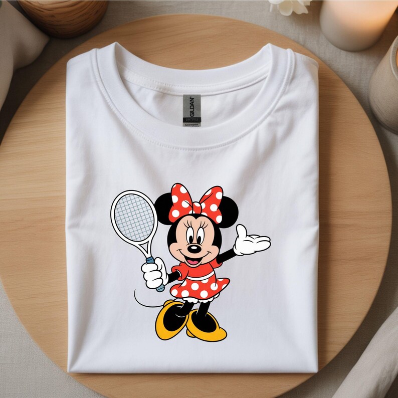 May include: White t-shirt featuring a cartoon Minnie Mouse character in a red dress with white polka dots, a matching bow, and a tennis racket. The character is in a tennis pose. The shirt is folded on a wooden surface.