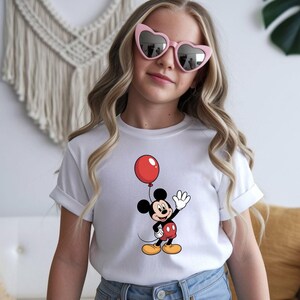 May include: White t-shirt featuring a cartoon Mickey Mouse holding a red balloon. Mickey Mouse is wearing red shorts, yellow shoes, and white gloves. The shirt has rolled-up sleeves.