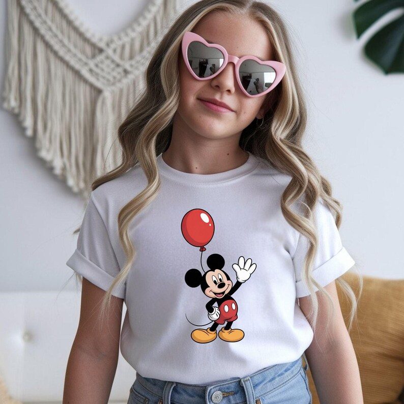 May include: White t-shirt featuring a cartoon Mickey Mouse holding a red balloon. Mickey Mouse is wearing red shorts, yellow shoes, and white gloves. The shirt has rolled-up sleeves.