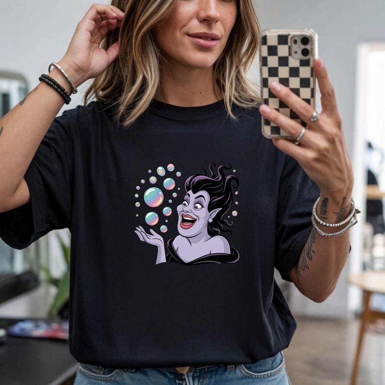 May include: Black t-shirt featuring a cartoon illustration of a purple-skinned character with black hair and a wide smile, surrounded by iridescent bubbles. The character is reaching out towards the bubbles. The shirt is a casual style.