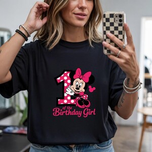 May include: Black t-shirt featuring a graphic of Minnie Mouse with a pink polka-dot number 1 and the text "of the Birthday Girl" in pink. The design includes a pink bow and Minnie Mouse's signature look.