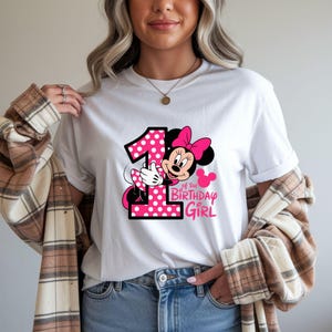May include: White t-shirt featuring a graphic of Minnie Mouse with a pink polka-dot number 1 and the text "of the Birthday Girl". Minnie Mouse is wearing a pink bow. The design is in pink and black.