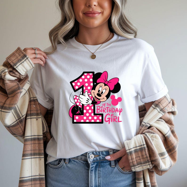 May include: White t-shirt featuring a graphic of Minnie Mouse with a pink polka-dot number 1 and the text "of the Birthday Girl". Minnie Mouse is wearing a pink bow. The design is in pink and black.
