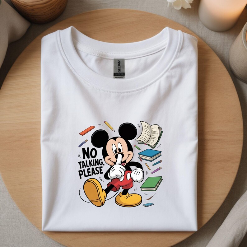 May include: White t-shirt with a Mickey Mouse graphic and the words "NO TALKING, PLEASE." The design includes books and pencils. The shirt is folded on a wooden surface.