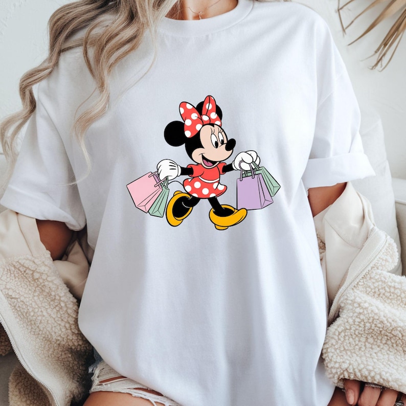 May include: White t-shirt featuring a cartoon Minnie Mouse character in a red dress with white polka dots and a matching bow. Minnie is carrying shopping bags in various colors. The shirt has short sleeves.