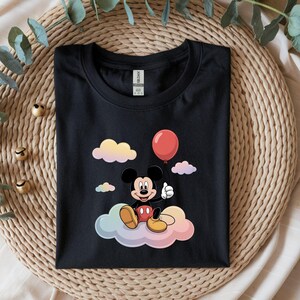 May include: Black t-shirt featuring a cartoon Mickey Mouse character sitting on a colorful cloud and holding a red balloon. The design includes pastel-colored clouds in the background. The shirt is folded neatly on a woven surface.