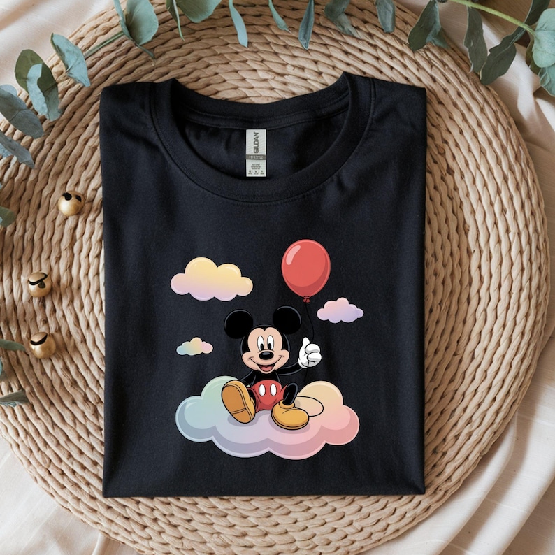 May include: Black t-shirt featuring a cartoon Mickey Mouse character sitting on a colorful cloud and holding a red balloon. The design includes pastel-colored clouds in the background. The shirt is folded neatly on a woven surface.