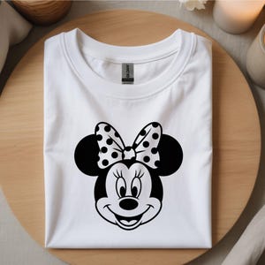 May include: White t-shirt featuring a black and white Minnie Mouse graphic. The graphic includes Minnie's face, large black ears, and a polka-dot bow. The shirt is folded neatly on a wooden surface.