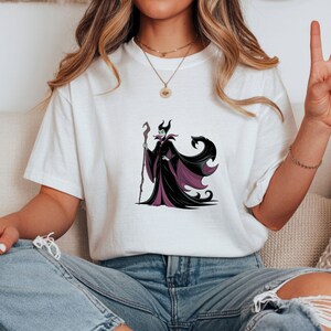 May include: White t-shirt featuring a graphic of a character in a black and purple gown, holding a staff. The character has a green face and black horns. The design is centered on the shirt.