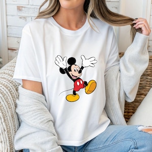 May include: White t-shirt featuring a cartoon image of Mickey Mouse. Mickey is depicted with black ears, a black body, red shorts, yellow shoes, and white gloves. The character is in a joyful pose with arms raised.