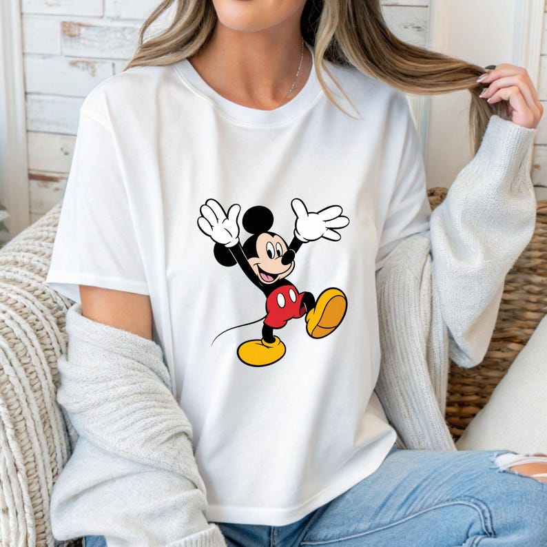 May include: White t-shirt featuring a cartoon image of Mickey Mouse. Mickey is depicted with black ears, a black body, red shorts, yellow shoes, and white gloves. The character is in a joyful pose with arms raised.