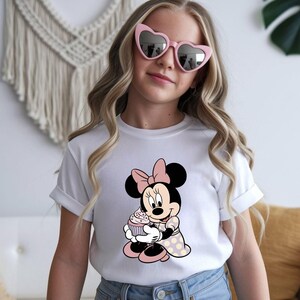 Op de afbeelding: Wit T-shirt met een Minnie Mouse-afbeelding die een cupcake vasthoudt. Minnie is afgebeeld in zwart, roze en wit, met een roze strik en een jurk met stippen. Het shirt heeft opgerolde mouwen.