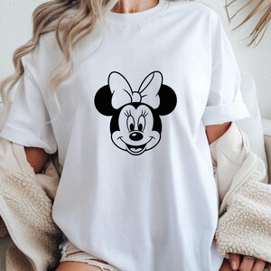 May include: White t-shirt featuring a black and white graphic of Minnie Mouse's face. The design includes her signature bow and smiling expression. The shirt is a casual style, suitable for everyday wear.