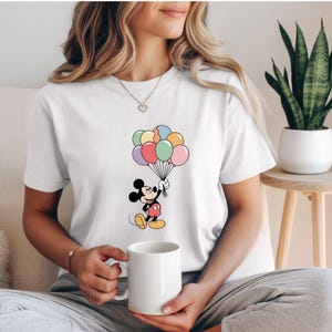 May include: White t-shirt featuring a cartoon image of Mickey Mouse holding a bunch of colorful balloons. Mickey is wearing red shorts and yellow shoes. The balloons are various pastel colors.
