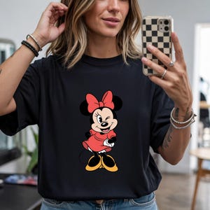 May include: Black t-shirt featuring a Minnie Mouse graphic. The cartoon character is depicted in a red dress and bow, with yellow shoes. The design is centered on the shirt.