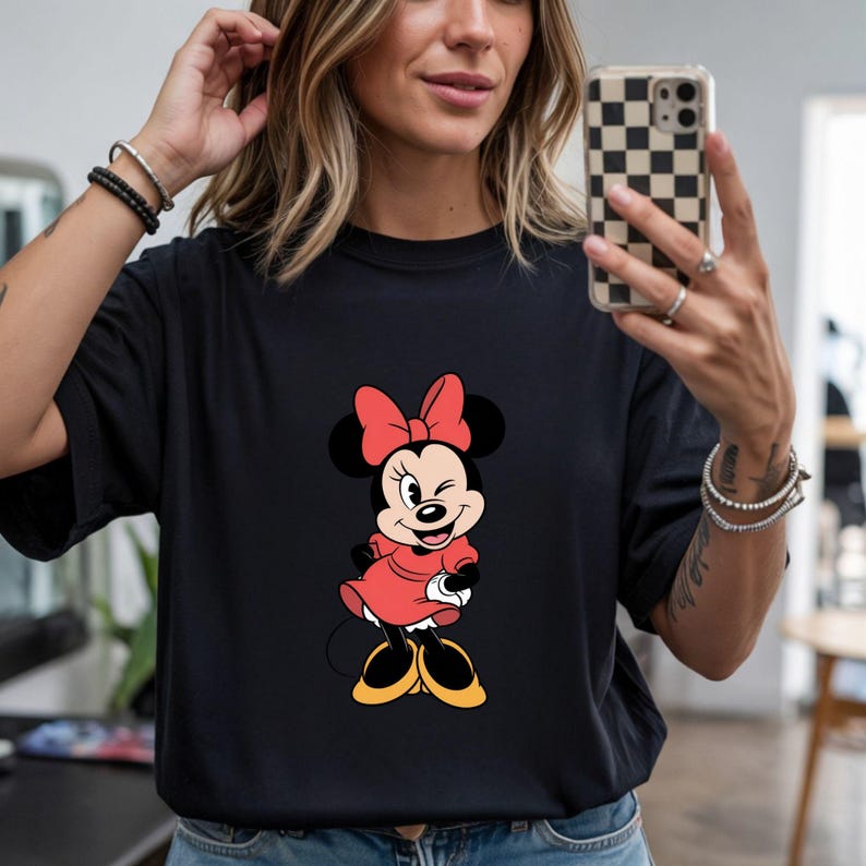 May include: Black t-shirt featuring a Minnie Mouse graphic. The cartoon character is depicted in a red dress and bow, with yellow shoes. The design is centered on the shirt.