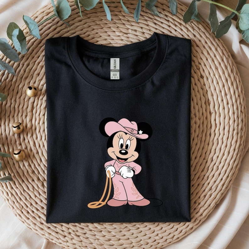 May include: Black t-shirt featuring a cartoon Minnie Mouse dressed as a cowgirl. Minnie is wearing a pink cowboy hat with a star, a pink outfit, and holding a lasso. The shirt is folded on a woven surface.
