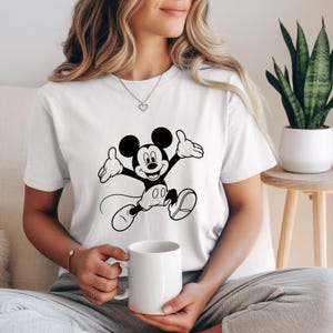 May include: White t-shirt featuring a black and white graphic of Mickey Mouse in a jumping pose. The cartoon character has a smiling expression and outstretched arms. The shirt is made of a soft material and is suitable for casual wear.