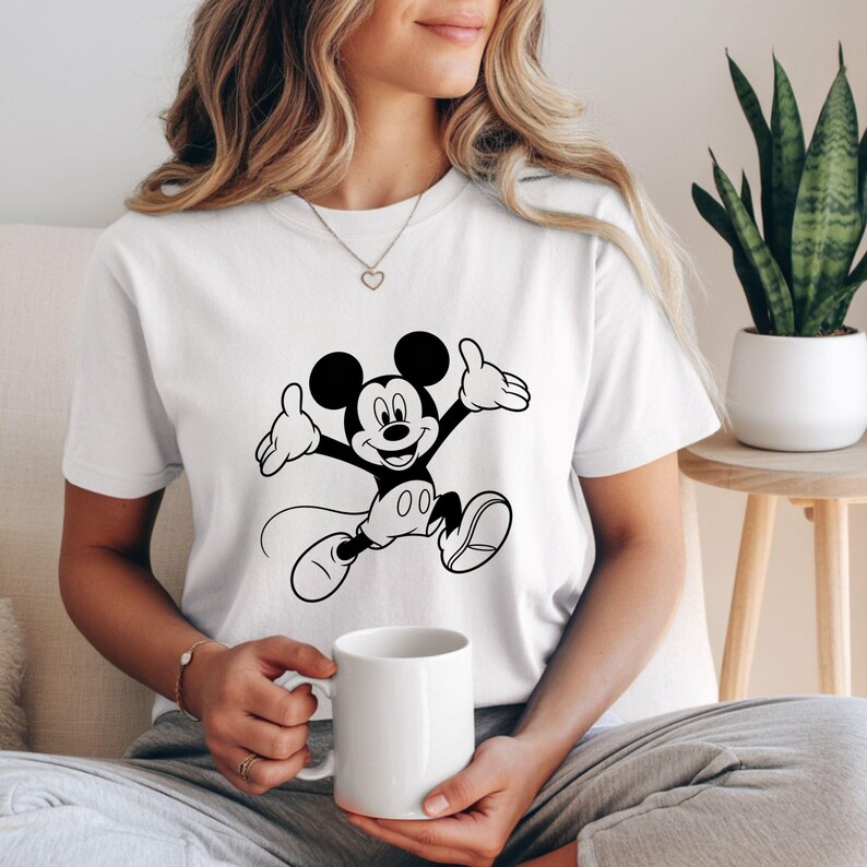 May include: White t-shirt featuring a black and white graphic of Mickey Mouse in a jumping pose. The cartoon character has a smiling expression and outstretched arms. The shirt is made of a soft material and is suitable for casual wear.
