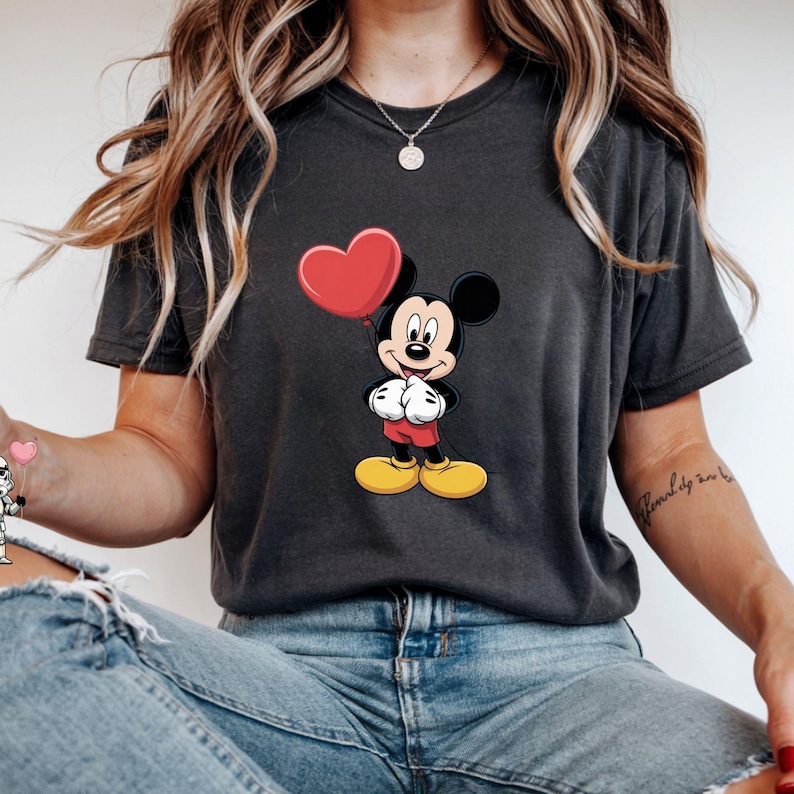 May include: Dark gray t-shirt featuring a cartoon Mickey Mouse holding a red heart-shaped balloon. Mickey is wearing red shorts, white gloves, and yellow shoes. The shirt is a casual style.