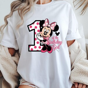 May include: White t-shirt featuring a Minnie Mouse graphic. The design includes a large number 1 with pink polka dots, Minnie Mouse, and the text "of the Birthday Girl" in pink script. The shirt has rolled-up sleeves.
