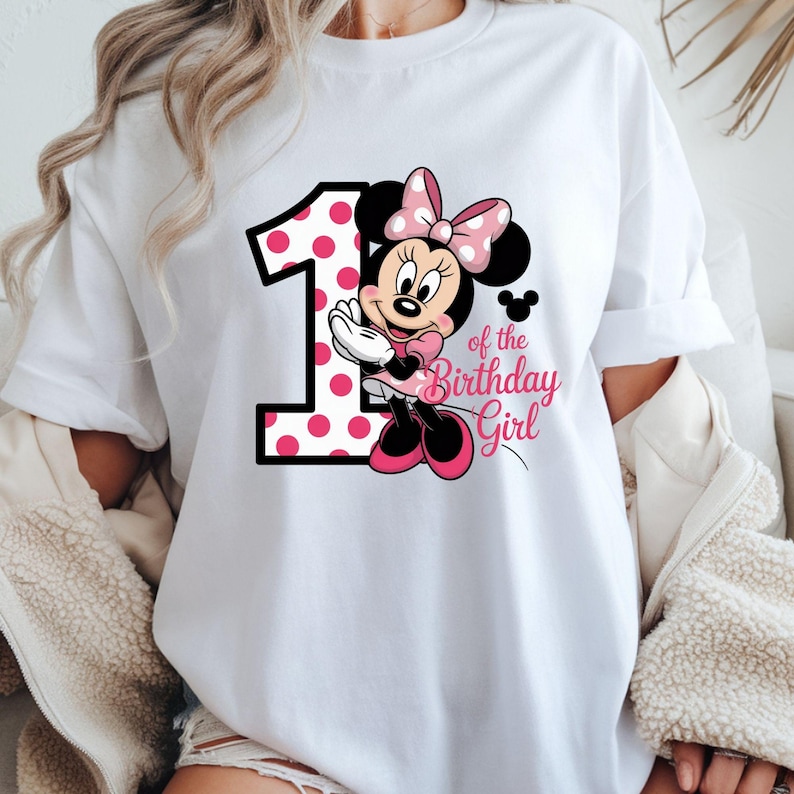 May include: White t-shirt featuring a Minnie Mouse graphic. The design includes a large number 1 with pink polka dots, Minnie Mouse, and the text "of the Birthday Girl" in pink script. The shirt has rolled-up sleeves.