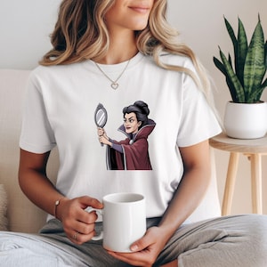 May include: White t-shirt featuring a cartoon illustration of a woman in a burgundy cape holding a mirror. The character has dark hair and is looking at the mirror. The t-shirt is a casual style.