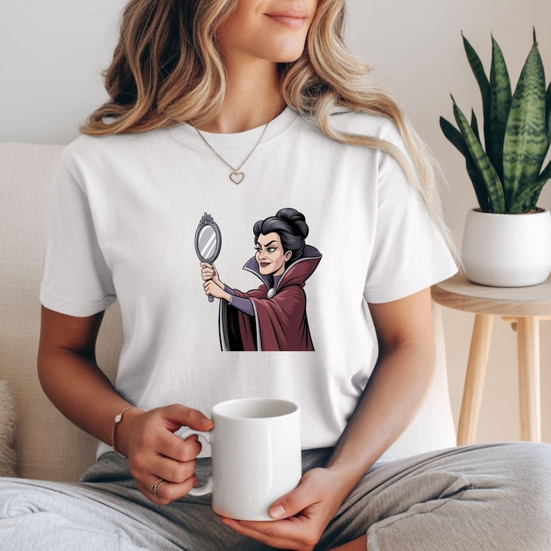May include: White t-shirt featuring a cartoon illustration of a woman in a burgundy cape holding a mirror. The character has dark hair and is looking at the mirror. The t-shirt is a casual style.